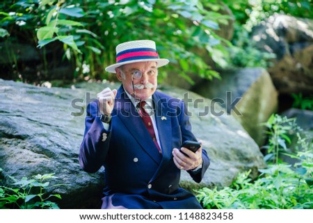 Portrait of an elderly, well-dressed senior gentleman in a double breasted suit, tie, hat and monocle smiling and doing a fist pump as he looks up from his smartphone. 