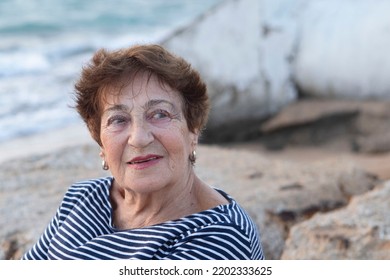 Portrait Of An Elderly Woman 80+ Relaxing On The Beach. Happy Granny, Pensioner With Light Make-up. Face With Wrinkles. Concept Of Pension, Good Quality Of Life, Health Insurance. Life Style. Sunset.