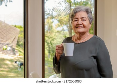 Portrait of an elderly woman drinking coffee in front of a window where you can see the backyard of her house.
