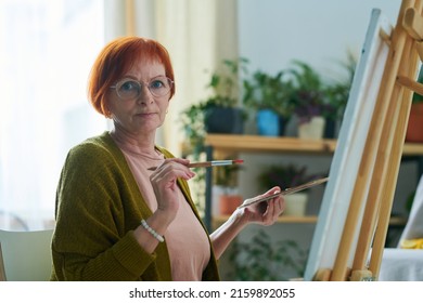 Portrait Of Elderly Woman In Eyeglasses Looking At Camera While Painting Picture On Canvas With Paints During Art Therapy At Home