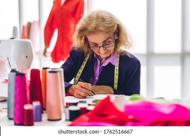 Portrait Of Elderly Woman Fashion Designer Stylish Sitting And Working With Sewing Machine.Attractive Senior Woman Working With Mannequins And Colorful Fabrics At Fashion Design Studio