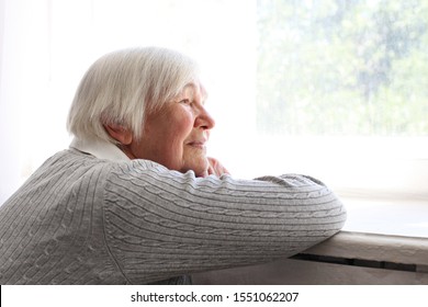 Portrait Of Elderly Woman With Grey Hair Sitting Alone By The Window. Old Lady Wearing A Cardigan Leaning On Windowsill By Herself, Expecting A Visit. Close Up, Copy Space, Background