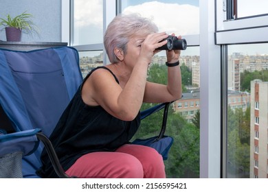 Portrait of an elderly woman looking through binoculars at other people's windows of houses sitting in an armchair. Concept: spying on neighbors, gathering information, espionage and gossip. - Powered by Shutterstock