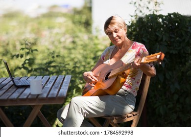Portrait of elderly woman playing  guitar and watching  online lessons  on laptop while practicing in the garden. Senior woman happy and enjoy life after retired. Online training