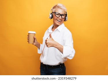 Portrait of elderly woman with short hair listening to music with headphones and holding takeaway coffee over yellow background. - Powered by Shutterstock