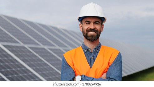 Portrait Of Electrician In Safety Helmet And Uniform On Background Field Of Photovoltaic Solar Panels. Male Technician At Solar Station.