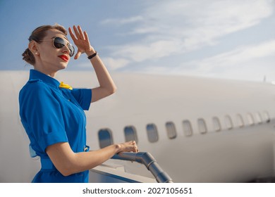 Portrait of elegant air stewardess in blue uniform and sunglasses holding hand near her head and looking far away, standing on airstair on a daytime. Aircrew, occupation concept - Shutterstock ID 2027105651