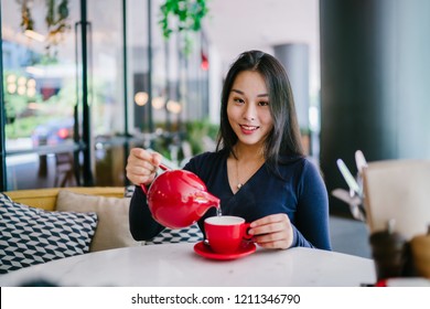 Portrait of an elegant Asian woman pouring hot tea from a red tea pot into her tea cup. She is young, attractive, beautiful and smiling as she pours her beverage. 