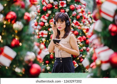 Portrait of a elegant and attractive middle-aged woman standing between a few decorated  Christmas trees during the day. She is smiling as she listens to music on her headphones and views the trees.