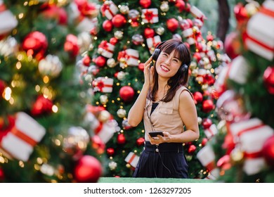 Portrait of a elegant and attractive middle-aged woman standing between a few decorated  Christmas trees during the day. She is smiling as she listens to music on her headphones and views the trees.