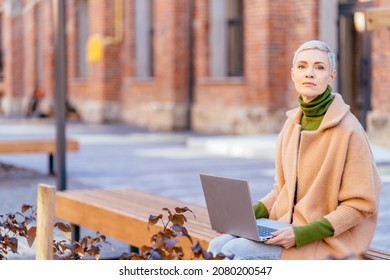 Portrait Of Elegant Blond Short Hair 40-45 Years Old Age Caucasian Trendy Woman Sitting On Benchwith Silver Laptop In Hands In Old Industrial Street With Red Brick Facades.