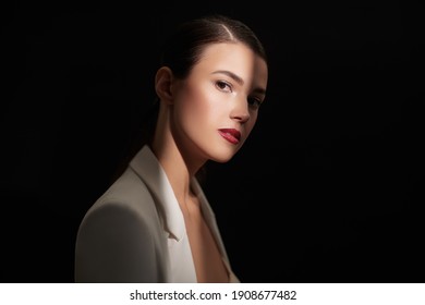 Portrait Of An Elegant Brunette Woman With Evening Makeup Standing In A Beam Of Light On A Black Background. Makeup And Cosmetics.