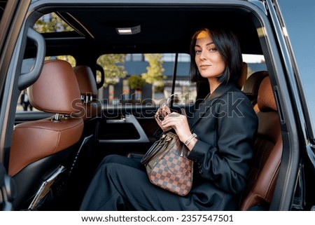 Portrait of an elegant business lady in black formal wear sitting on backseat of luxury SUV vehicle. Concept of business trips and transportation