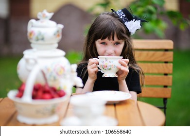 Portrait Of Elegant Child Girl In A Black Dress Having A Tea Party Outdoors, Focus On The Background With Girl's Face