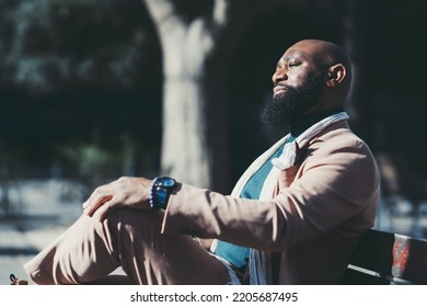 A Portrait Of An Elegant Confident Bald Black Man With A Well-groomed Beard Enjoying The Sun While Sitting On A Park Bench; A Side View Of A Bearded African Man In A Tailored Suit, Sitting Outdoors