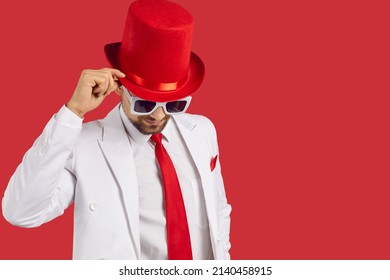 Portrait of elegant gentleman in white suit and red top hat. Handsome young man wearing jacket, tie, tophat and glasses makes a bow standing on red colour text copy space fashion studio background