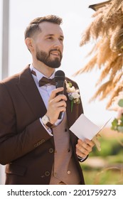 Portrait of elegant groom in wedding brown suit with boutonniere and bowtie, holding paper with oath and microphone while standing on outdoors engagement ceremony - Shutterstock ID 2201759273