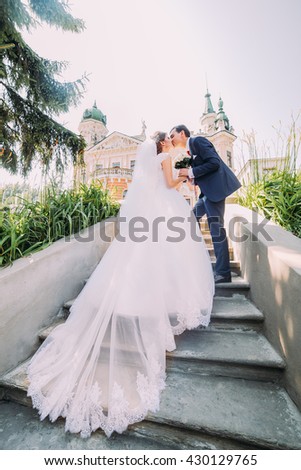 Portrait of elegant stylish young wedding couple kissing on stairs in park. Romantic antique palace at background