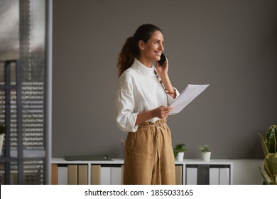Portrait of elegant successful businesswoman speaking by smartphone and smiling happily while standing against grey wall in office, copy space