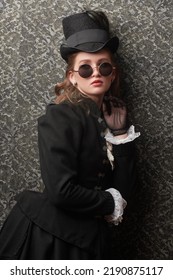 Portrait Of An Elegant Victorian Woman In Traditional 19th Century Clothes And Stylish Sunglasses Posing On A Vintage Background. Style Of The Late 19th - Early 20th Century.