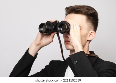 Portrait Of Elegant Young Man Wearing Black  Suit Looking Through Binoculars. Studio Shot On Gray Background