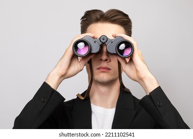 Portrait Of Elegant Young Man Wearing Black  Suit Looking Through Binoculars. Studio Shot On Gray Background