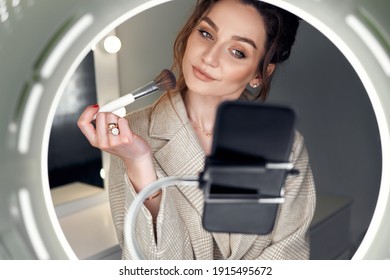 Portrait of an elegant young woman blogger stands in front of the phone and powders her face. She looks into the camera with a smile while recording a video. - Powered by Shutterstock