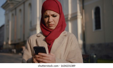 Portrait Of Embarassed Young Muslim Woman Looking At Smartphone. Surprised Arab Girl On The Busy Street. Shocking News.