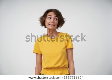 Portrait of embarrassed young female lady with short brown hair looking aside and showing teeth confusedly, keeping hands along her body while posing over white background