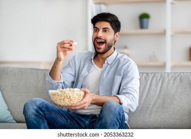 Portrait Of Emotional Arab Man Watching TV And Eating Popcorn, Resting On Sofa At Home, Free Space. Excited Guy Enjoying Good Movie Or Program On Weekend