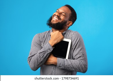 Portrait of emotional black guy hugging tablet with blank screen, holding it tight near chest, blue studio wall