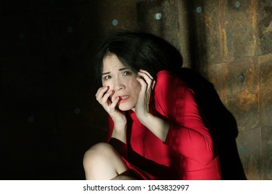 Portrait Of An Emotional And Frightened Young Woman With Loose Black Hair And A Red Dress, Looking With Fear In The Window, Pressing Her Hands To Her Face Against The Background Of A Rusty Metal Wall