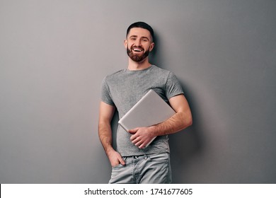 Portrait Of Emotional Handsome Guy Holding Laptop Leaning Against Gray Wall. Look At The Camera And Laugh.