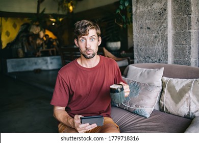 Portrait Of Emotional Male Millennial Guy Confused And Surprised Downloading Interesting Book On E-reader, Young 20s Male Looking At Camera Confused With Something Holding Mug And Digital Tablet