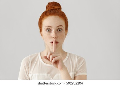 Portrait of emotional redhead girl with freckles and hair bun holding index finger at her lips, saying 'shh', asking for silent or to keep her secret. Human face expressions, emotions and feelings