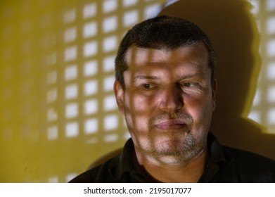 Portrait Of An Emotionless Adult Male Against Yellow Wall. Pattern Of Repeating Square Patches Of Light On Wall And Man's Face. A Man With A Serene Expression On His Face. People, Emotion Concept.