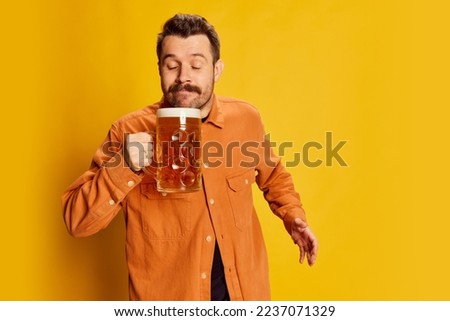 Portrait of emotive handsome man in orange shirt posing with lager foamy beer glass isolated over yellow background. Delightful look. Emotions, beer degustation, facial expression, Oktoberfest concept