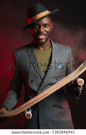 Portrait of energetic, sporty, self-assured handsome african man infashionable blazer suit holding longboard in hands, posing isolated in studio on dark background. Sport, Health anf Active lifestyle