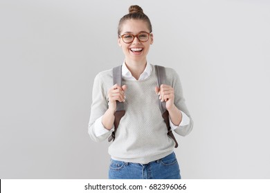 Portrait of energetic teenager girl isolated on gray background pulling forward straps of gray hipster backpack, laughing happily as if anticipating friendly meeting or great leisure time in open air.
