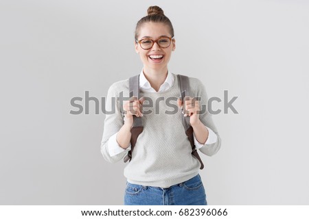 Portrait of energetic teenager girl isolated on gray background pulling forward straps of gray hipster backpack, laughing happily as if anticipating friendly meeting or great leisure time in open air.