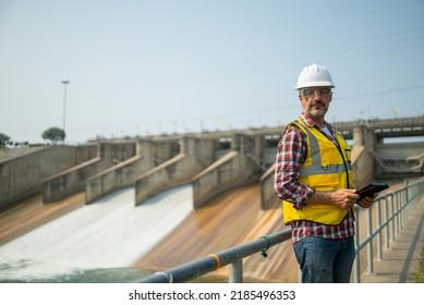 Portrait Of Engineer Wearing Yellow Vest And White Helmet With Tablet Working Day On A Water Dam With A Hydroelectric Power Plant. Renewable Energy Systems, Sustainable Energy Concept
