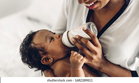 Portrait Of Enjoy Happy Love Family African American Mother Playing With Adorable Little African American Baby.Mom Feeding Bottle Of Milk To Baby Cute Son In A White  Bedroom.Love Of Black Family 