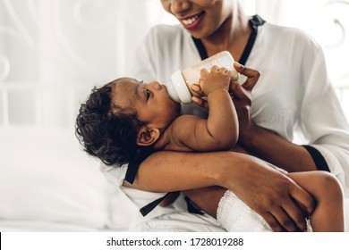 Portrait Of Enjoy Happy Love Family African American Mother Playing With Adorable Little African American Baby.Mom Feeding Bottle Of Milk To Baby Cute Son In A White  Bedroom.Love Of Black Family 