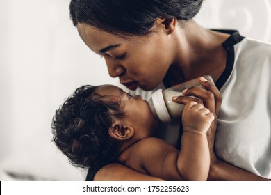 Portrait Of Enjoy Happy Love Family African American Mother Playing With Adorable Little African American Baby.Mom Feeding Bottle Of Milk To Baby Cute Son In A White  Bedroom.Love Of Black Family