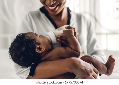 Portrait Of Enjoy Happy Love Family African American Mother Playing With Adorable Little African American Baby.Mom Feeding Bottle Of Milk To Baby Cute Son In A White  Bedroom.Love Of Black Family 