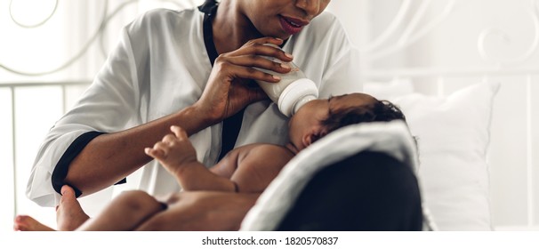 Portrait Of Enjoy Happy Love Family African American Mother Playing With Adorable Little African American Baby.Mom Feeding Bottle Of Milk To Baby Cute Son In A White  Bedroom.Love Of Black Family 