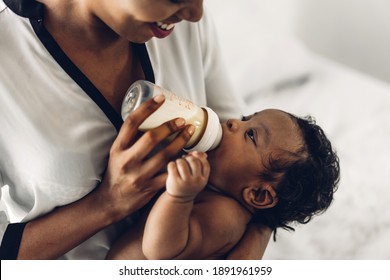 Portrait Of Enjoy Happy Love Family African American Mother Playing With Adorable Little African American Baby.Mom Feeding Bottle Of Milk To Baby Cute Son In A White  Bedroom.Love Of Black Family 