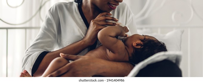 Portrait Of Enjoy Happy Love Family African American Mother Playing With Adorable Little African American Baby.Mom Feeding Bottle Of Milk To Baby Cute Son In A White Bedroom.Love Of Black Family