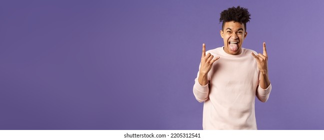 Portrait of enthusiastic, excited funny young man having fun, enjoying awesome rock concert, show heavy metal sign, stick tongue and rejoicing hear favorite band on stage, purple background - Shutterstock ID 2205312541