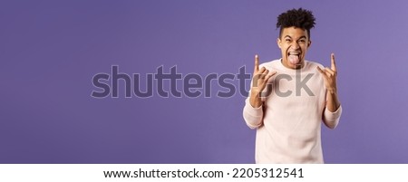 Portrait of enthusiastic, excited funny young man having fun, enjoying awesome rock concert, show heavy metal sign, stick tongue and rejoicing hear favorite band on stage, purple background